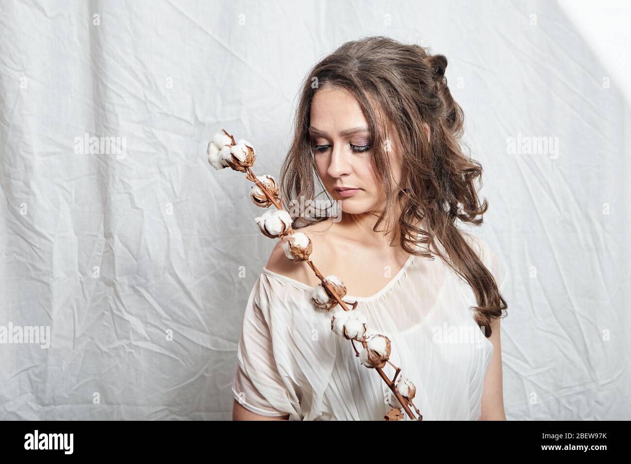 Portrait of young woman with cotton plant with fluffy flowers on white background. Attractive lady with long wavy brown hair Stock Photo