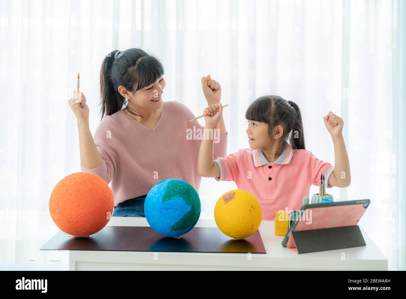 Asian elementary schoolgirl with mother painting the moon in science class learning about the solar system via video conference with teacher and other Stock Photo