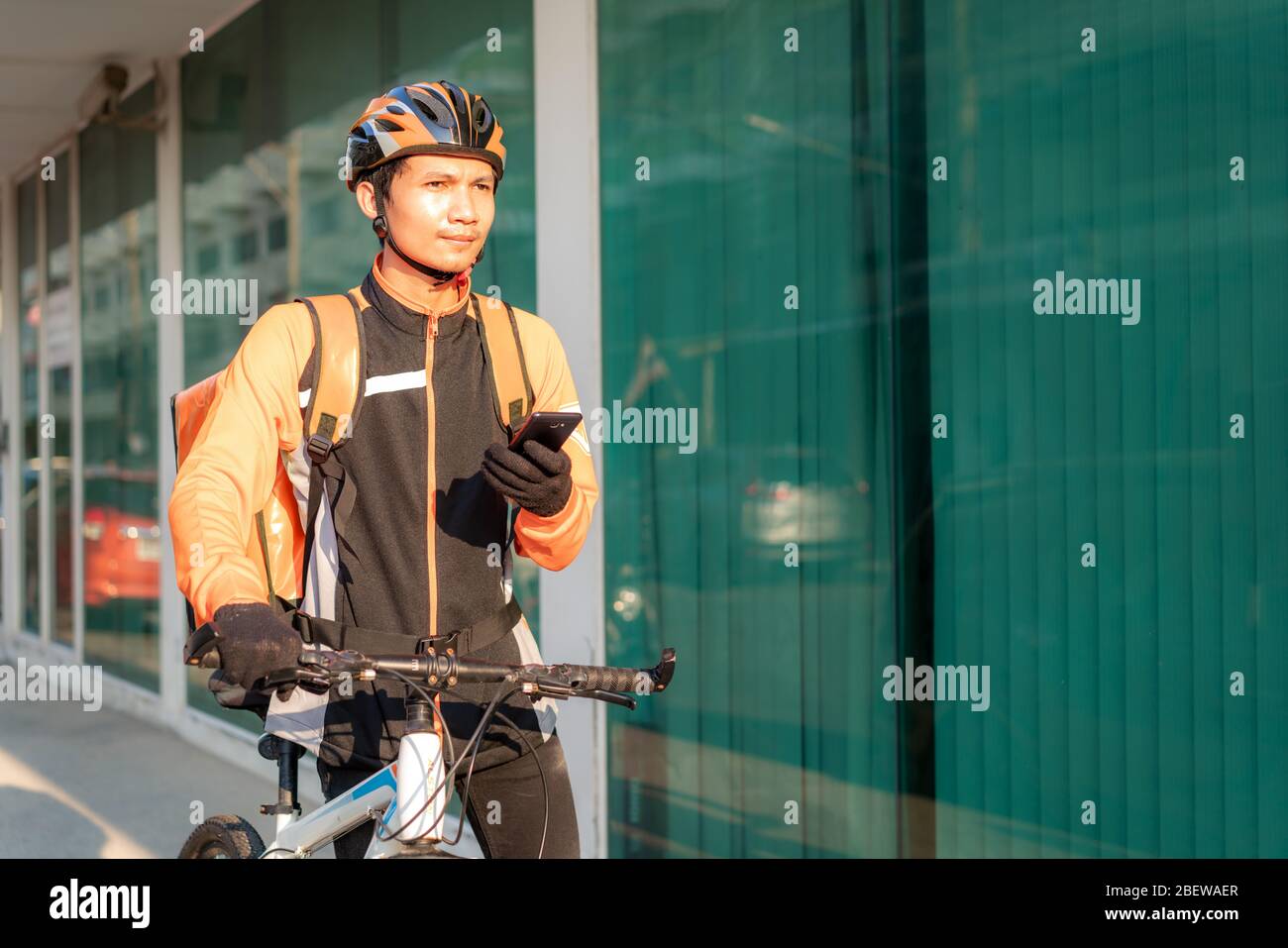 Asian man courier checking customer address in map in smartphone on bicycle delivering food in town streets with a hot food delivery from take aways a Stock Photo