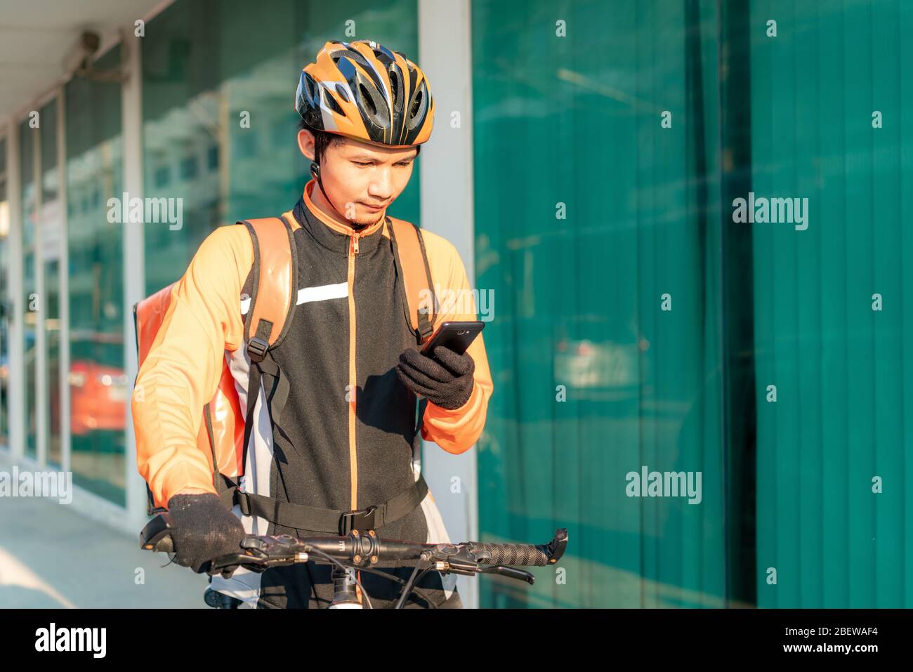 Asian man courier checking customer address in map in smartphone on bicycle delivering food in town streets with a hot food delivery from take aways a Stock Photo
