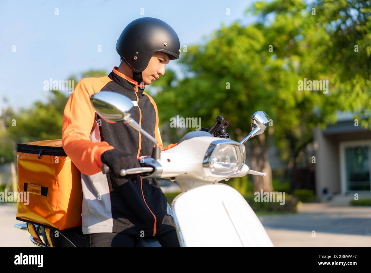 Asian man courier checking customer location in map in smartphone on scooter delivering food in town streets with a hot food delivery from take aways Stock Photo