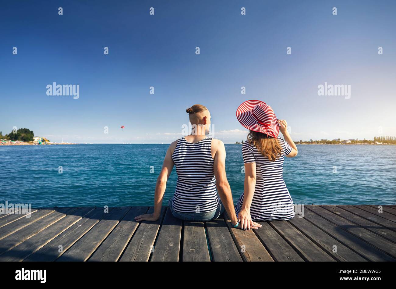 Romantic couple in striped dress sitting on pier and looking at blue Lake Issyk Kul in Kyrgyzstan. Summer and Sea concept. Stock Photo