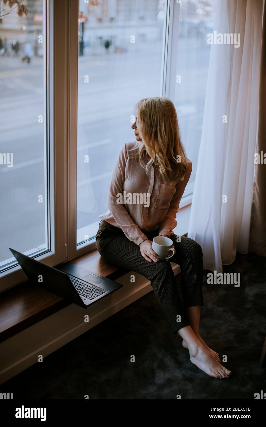 Pretty young woman working on laptop in living room on the window sill ...