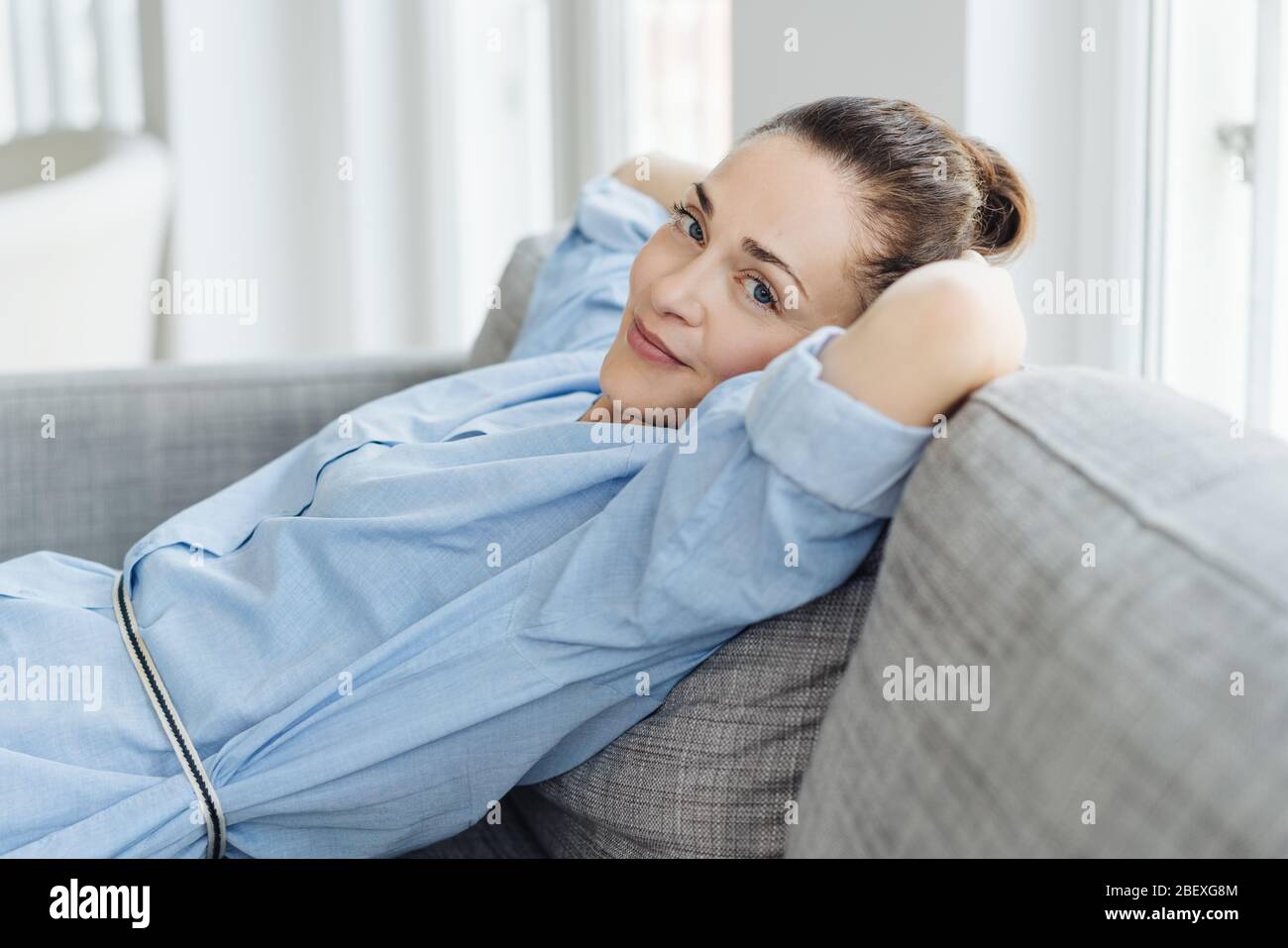 Trendy relaxed young woman unwinding at home on a comfortable sofa turning to smile at camera Stock Photo