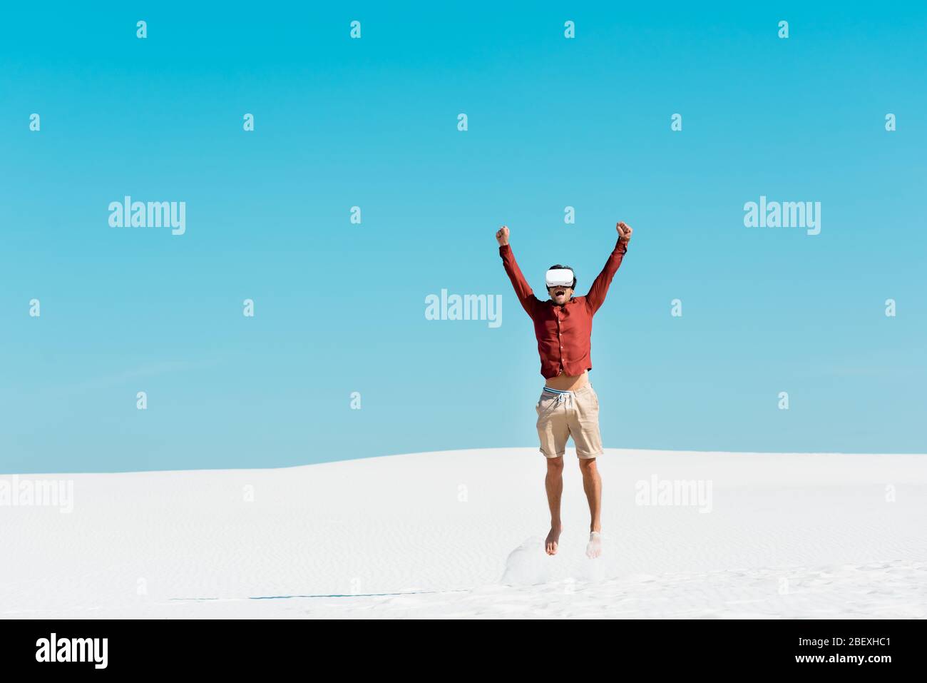 man on sandy beach in vr headset jumping against clear blue sky Stock Photo
