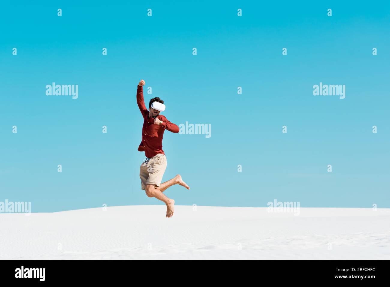 man on sandy beach in vr headset jumping against clear blue sky Stock Photo