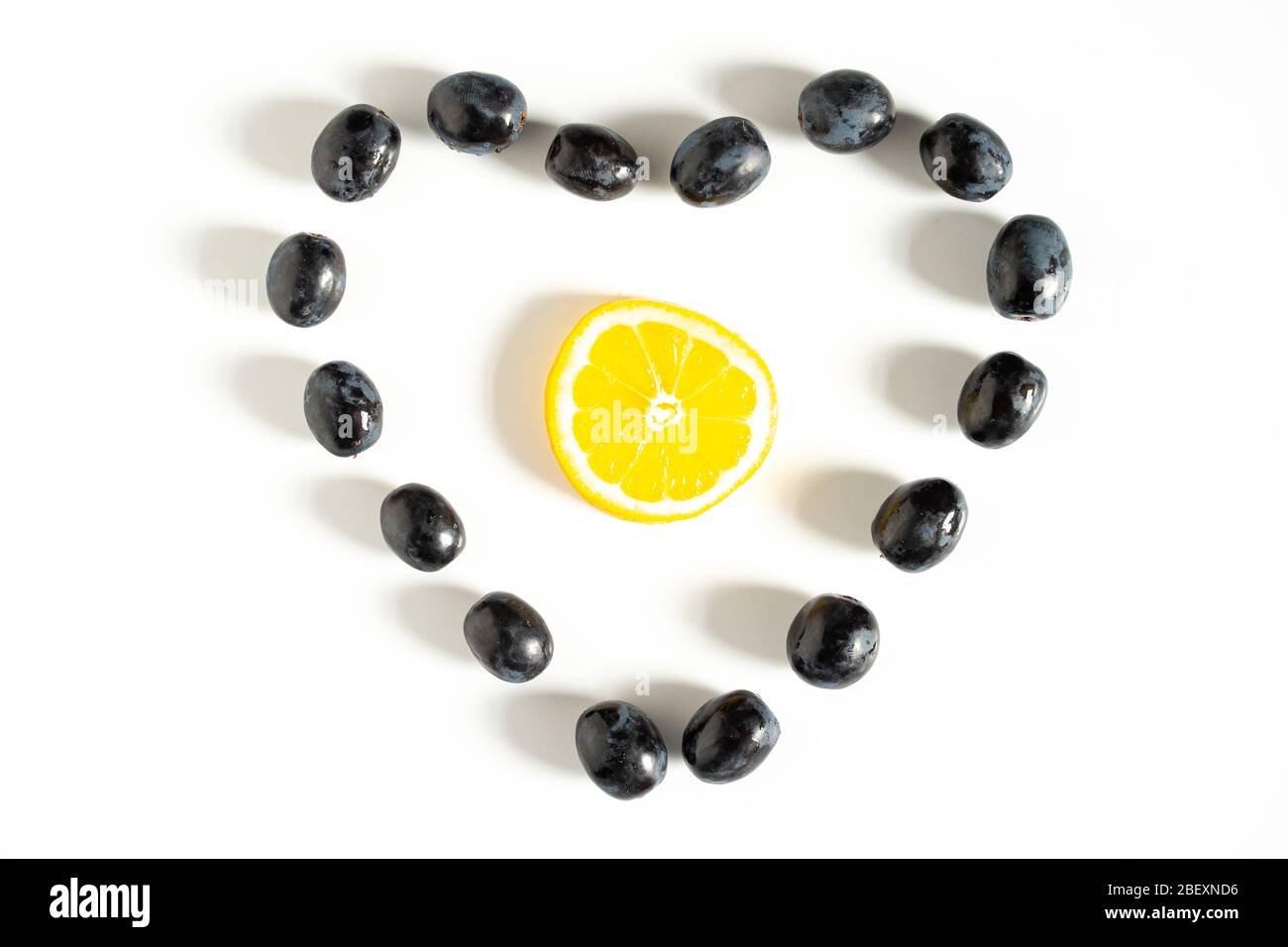 Black grapes laid out in the shape of a love heart with a lemon slice in the center against a plain white background Stock Photo