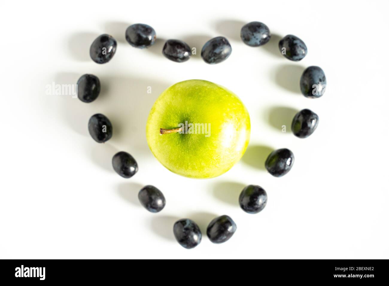 Black grapes laid out in the shape of a love heart with a granny smith apple in the center against a plain white background Stock Photo