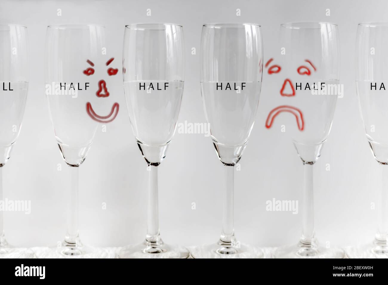 Six wine glasses, half full with drawn sad and happy faces in background depicting concept of glass half full, or glass half empty. Stock Photo
