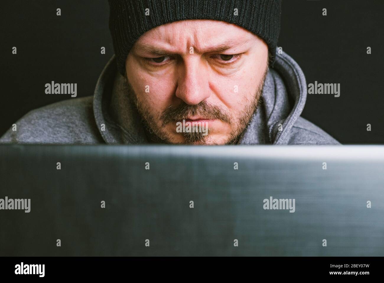 Man wearing black cap and hoodie sitting behind the computer monitor in the dark, close up, hacking computer system concept Stock Photo