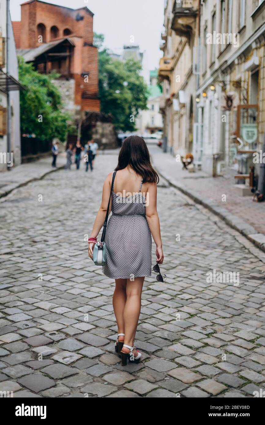Young woman in a gray sundress on a city tour. Brunette woman in dress and sandals walking on city streets. Girl in a handbag and glasses Stock Photo