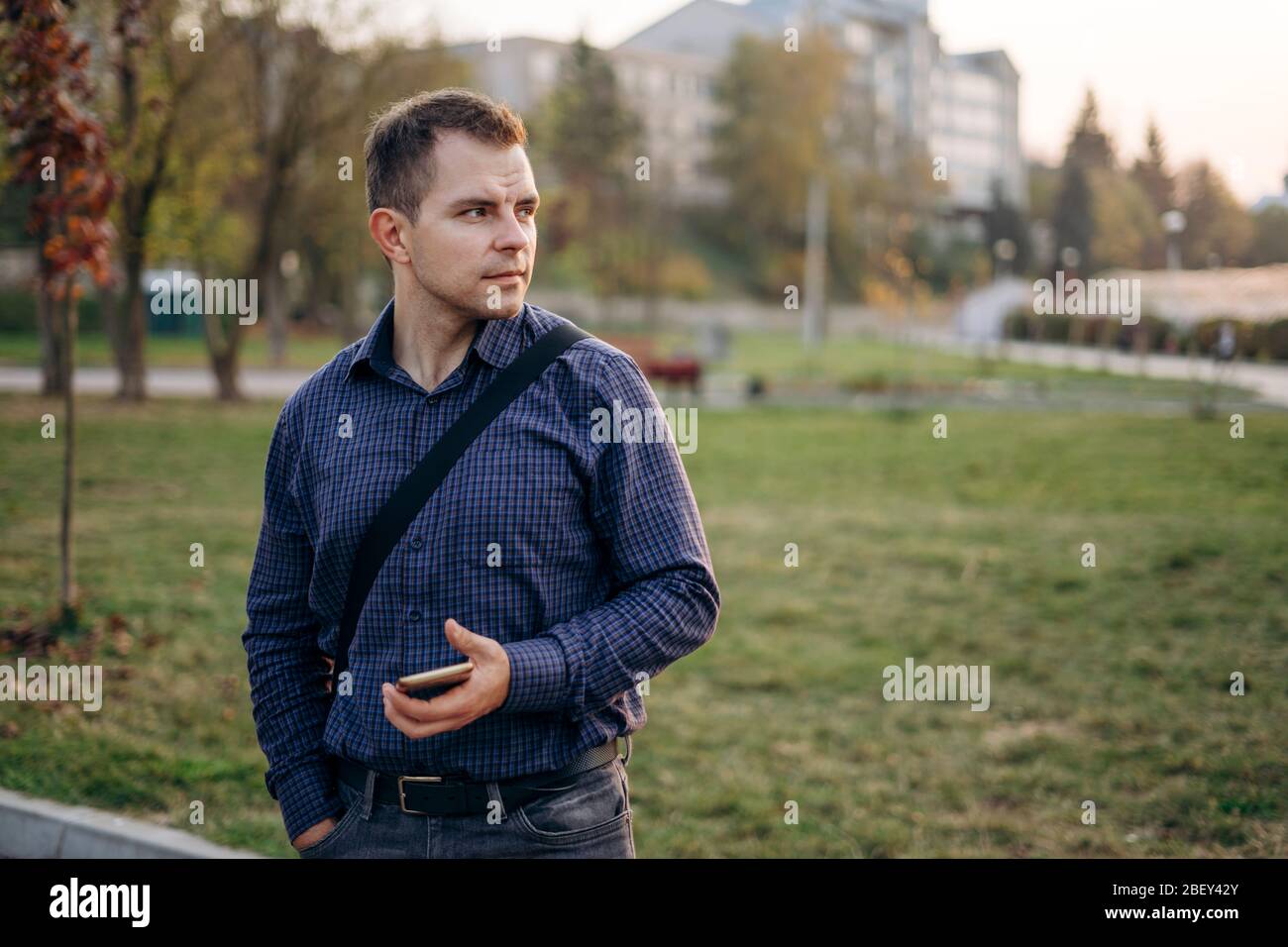 A man in a blue shirt stands in the park. A man holds his phone and walks in the park Stock Photo