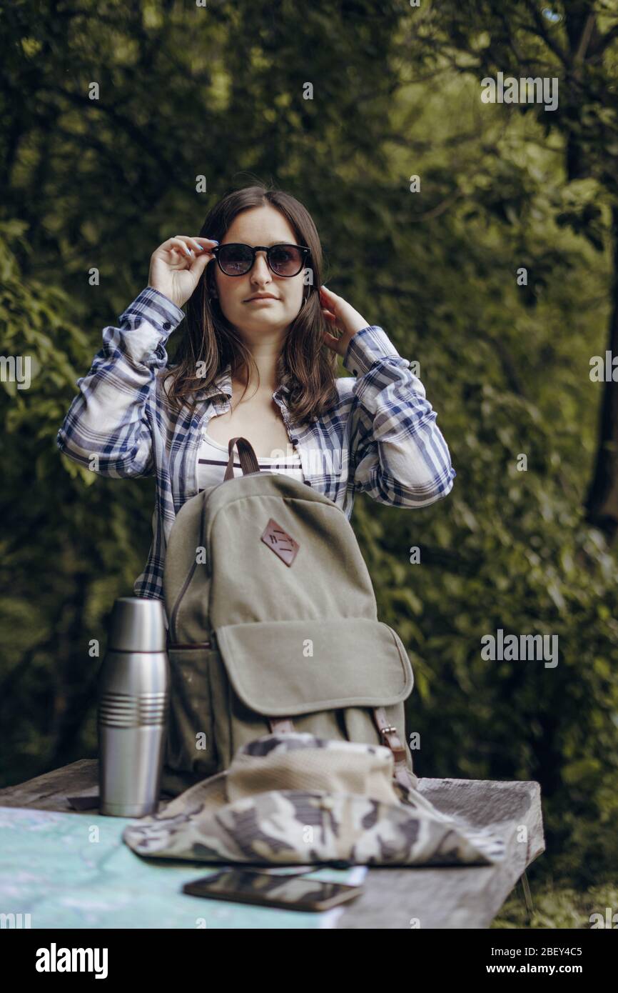 Girl in sunglasses in the forest with a backpack. Tourist map, thermos on a wooden table. Stock Photo