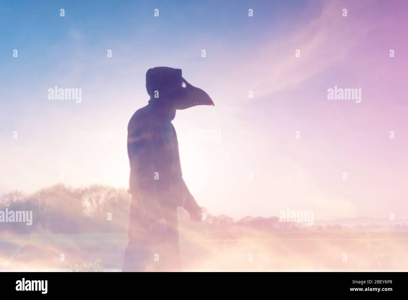 A double exposure of a moody, hooded figure with a plague doctor mask, silhouetted against the sunset. With an abstract, experimental dream like edit. Stock Photo