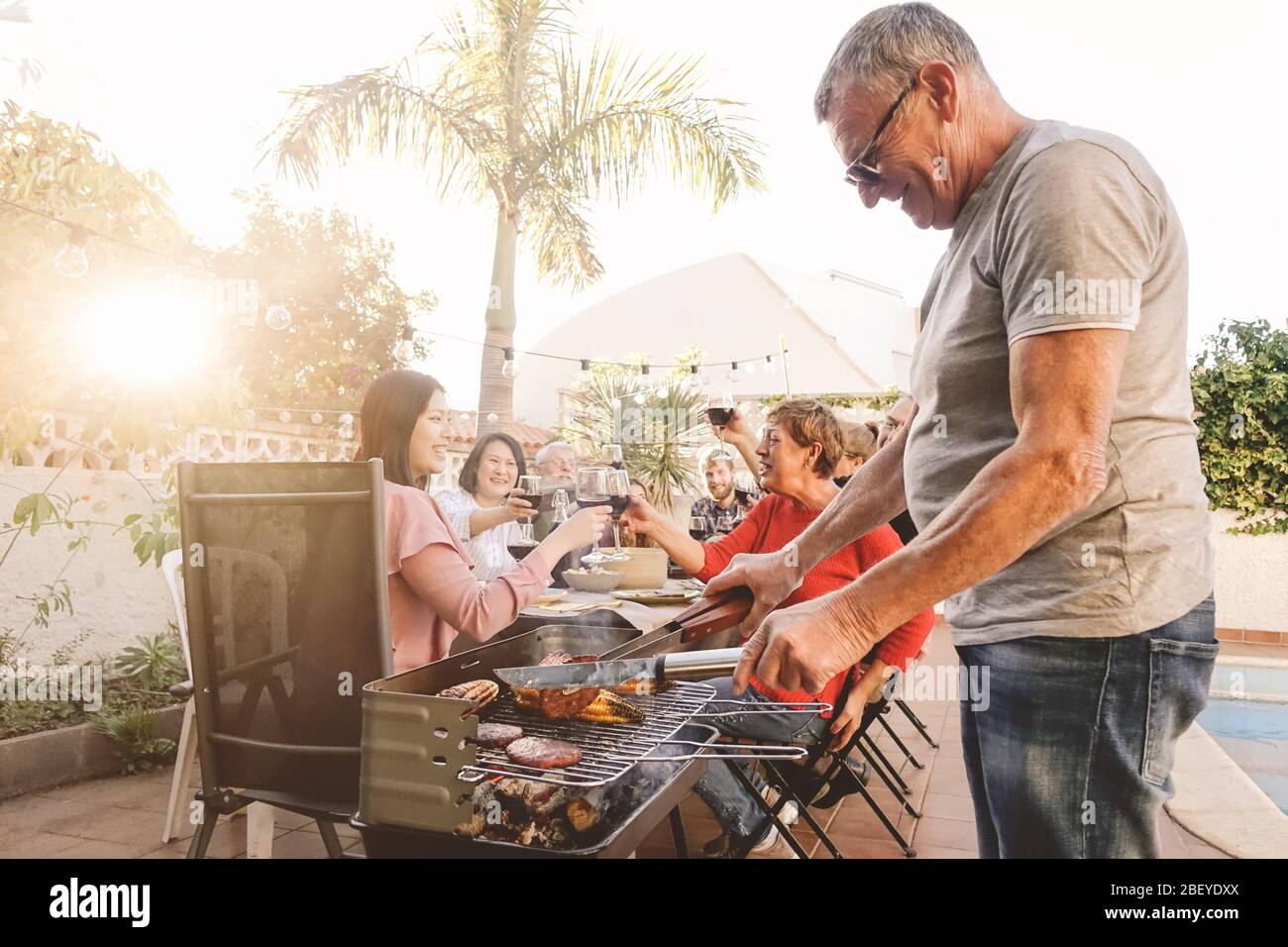 Happy family cheering and toasting with red wine in barbecue party - Chef senior man grilling meat and having fun with parents Stock Photo