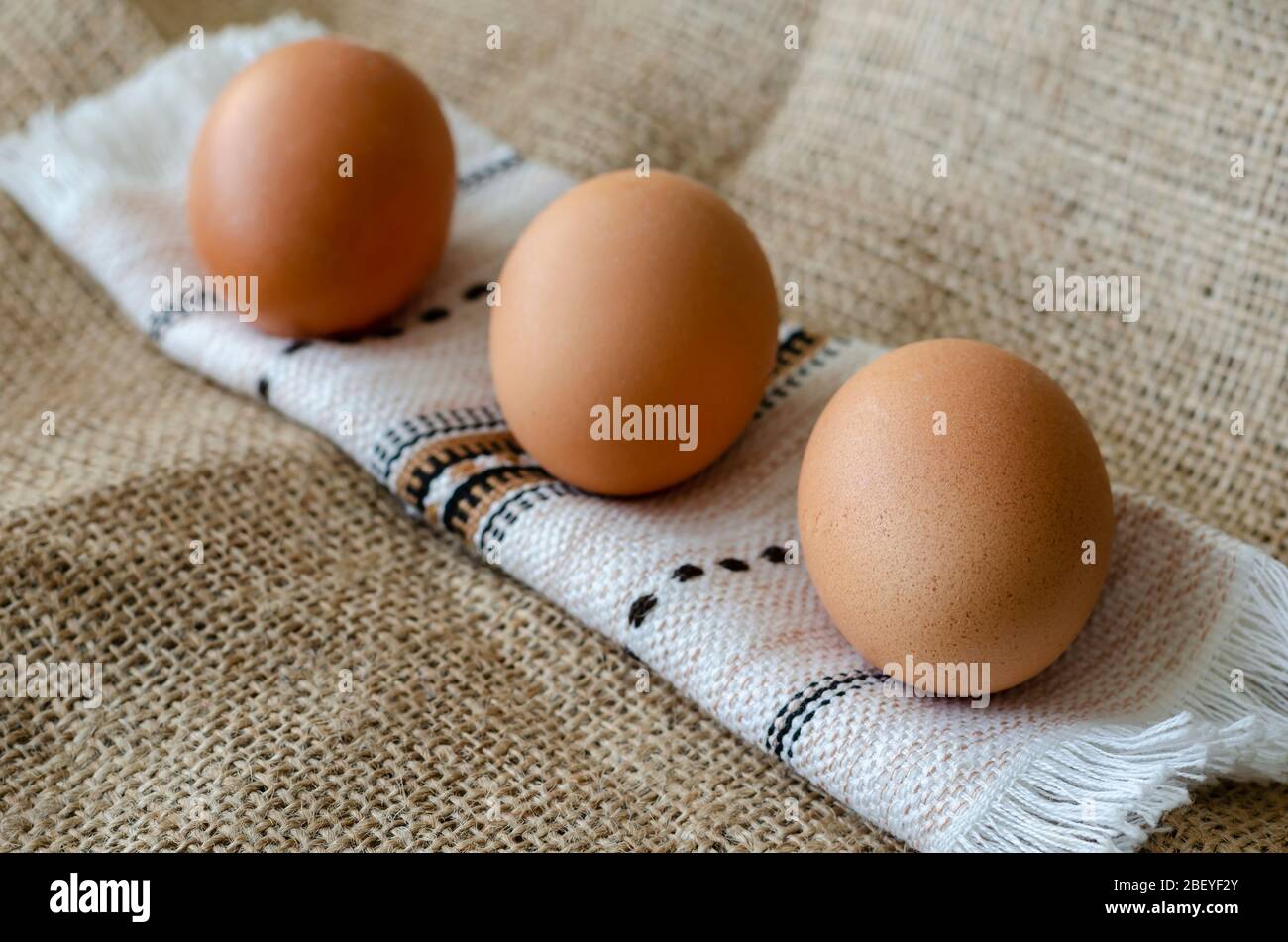 Three brown chicken eggs on burlap. A line of raw farm eggs on a rushnyk. A rolled towel with eggs on burlap. Angled side view. Close-up. Selective fo Stock Photo