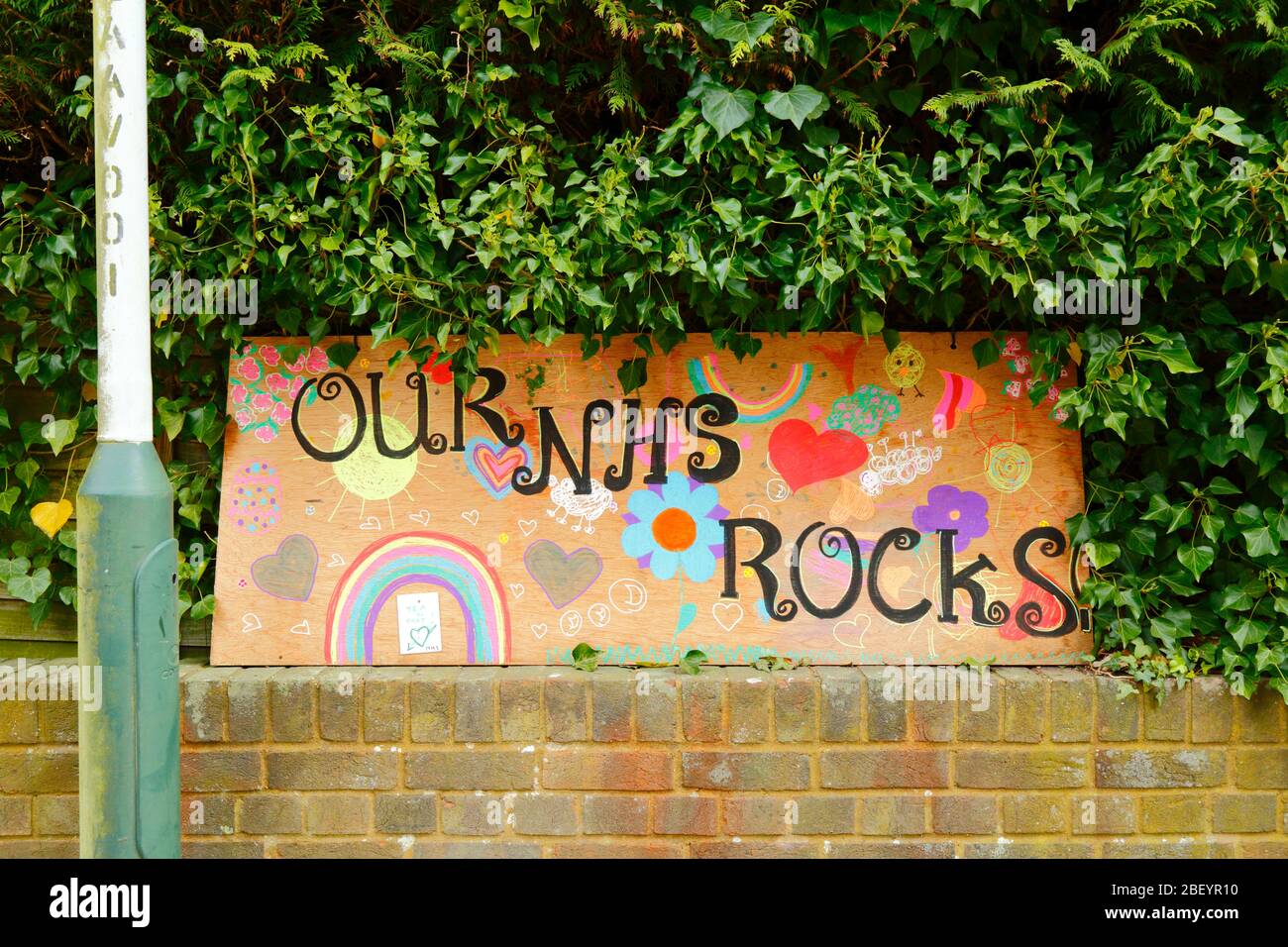 16th April 2020, Southborough, Kent, UK: Child's drawings of a rainbow, hearts and Our NHS Rocks message for NHS staff and key workers on a plywood board outside a hedge in residential area during the government imposed quarantine / lockdown to reduce the spread of the coronavirus. Children across the country have been putting drawings of rainbows in windows to spread hope and encourage people to stay cheerful during the pandemic. Stock Photo