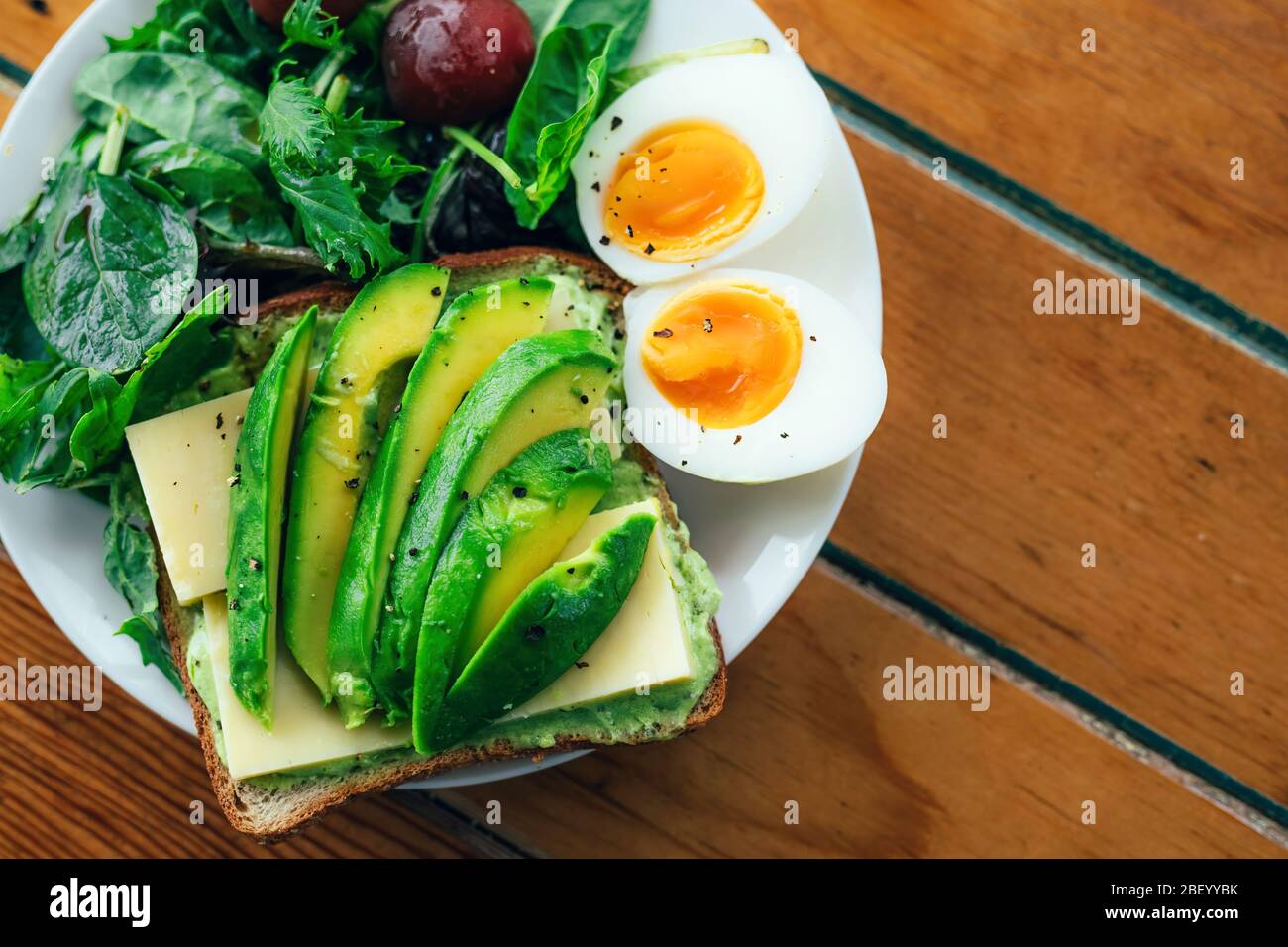 Toast with avocado, boiled egg, spinach and tomatoes on white plate with knife and fork on serviette and rustic wooden background. Stock Photo