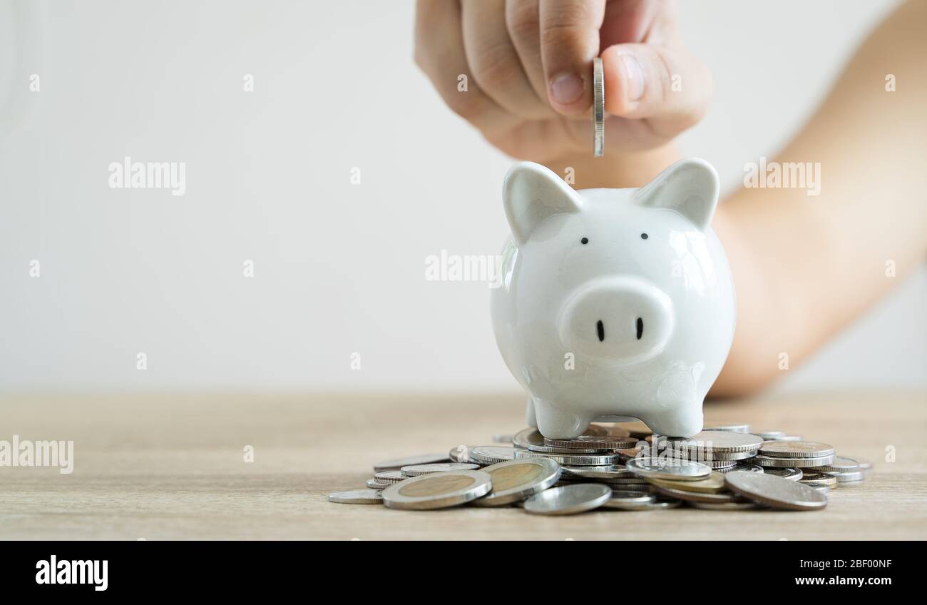Money savings concepts hand holding coin to put in piggy bank to spend on expenses such as savings, tourism, investment, emergency, retirement on wood Stock Photo