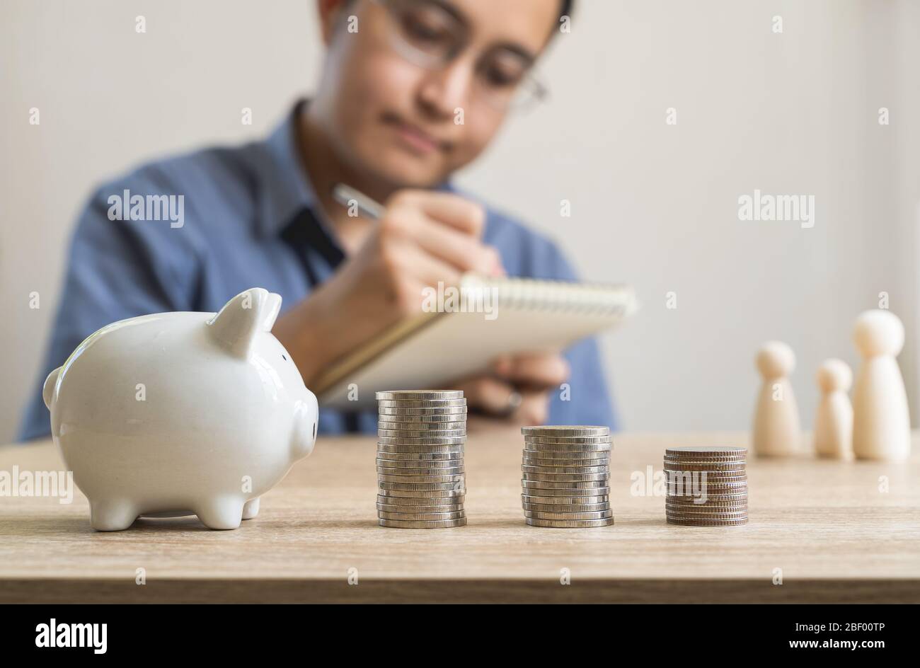 Money savings concepts  piggy bank and stack coins with blur man who's taking notes about saving money for family and family wooden dolls.on wooden ta Stock Photo