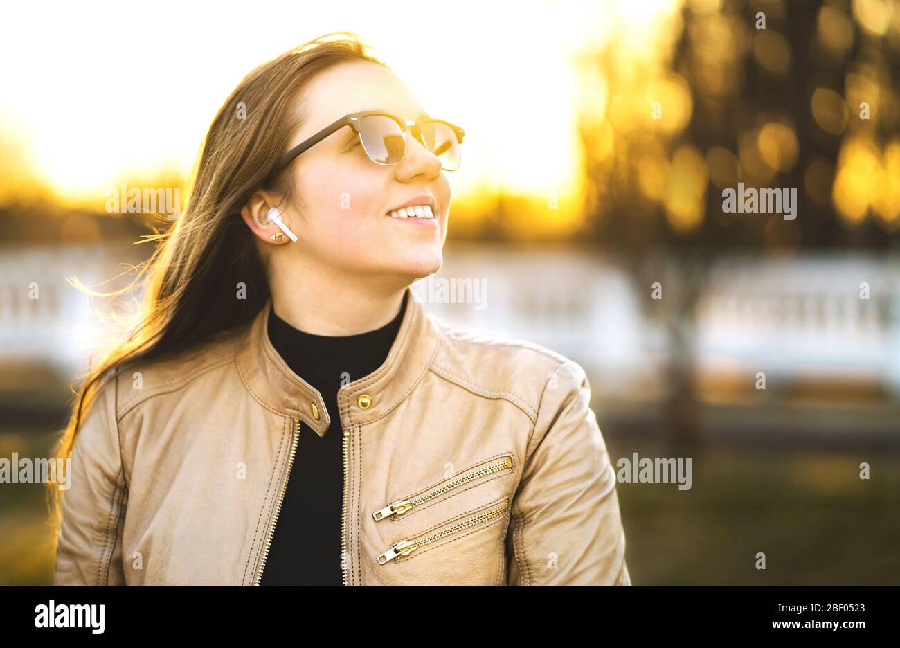 Wireless headphones, earbuds. Woman listening to music with earpods in a park. Happy young lady smiling. Sunglasses and leather jacket. Stock Photo