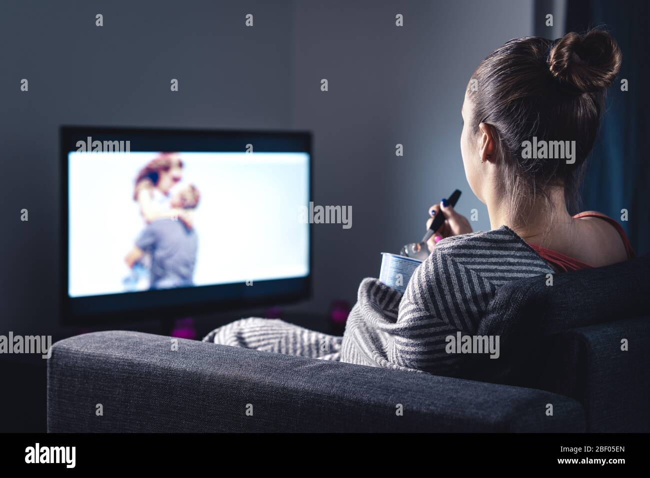 Woman watching romantic movie and eating ice cream. Sad lonely single girl streaming series or film on tv home at night. Person with stream on tv. Stock Photo