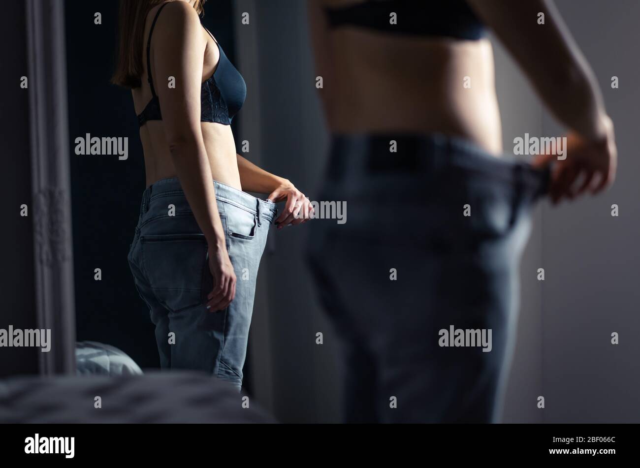 Skinny slim woman wearing big large pants in front of a mirror after weight loss and diet. Underweight girl with unhealthy stress of being fat. Stock Photo