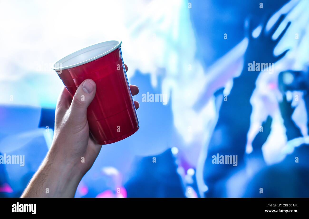 Red party cup in hand in night club, bar or college student event. Plastic beer mug. People having fun in blue nightclub disco lights on dance floor. Stock Photo