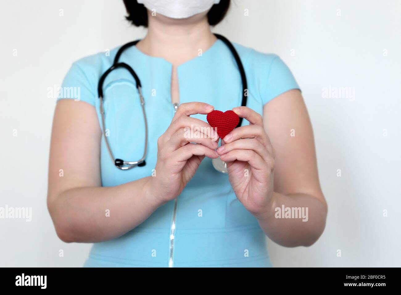 Saving lives during a covid-19 coronavirus pandemic, woman doctor in medical mask holding red knitted heart in hands. Concept of cardiologist Stock Photo