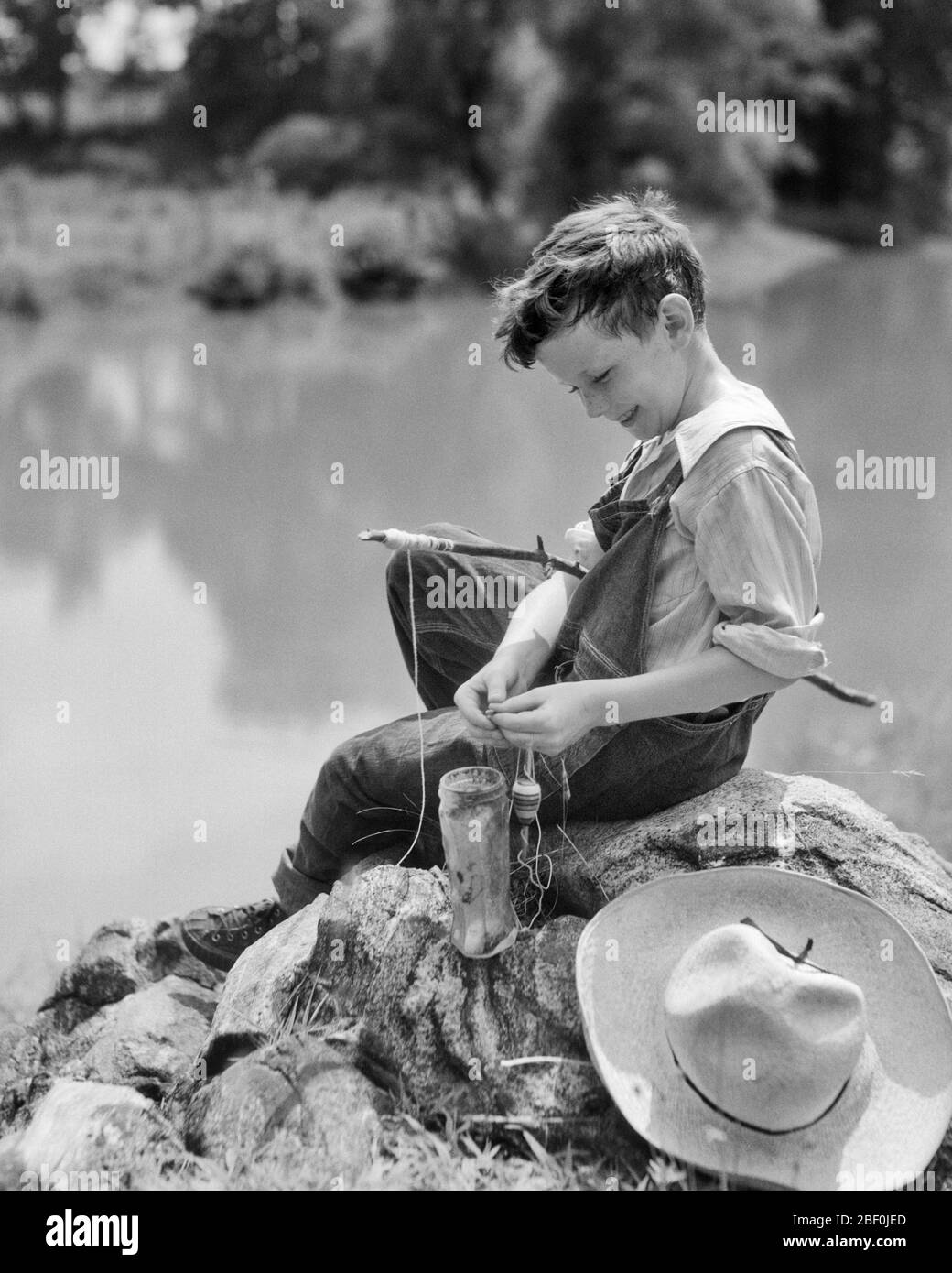 1930s SMILING YOUNG BOY SITTING ON ROCK BY STREAM PUTTING BAIT ON HOOK AND  LINE OF FISHING ROD MADE FROM A STICK - a2490 HAR001 HARS ROD DENIM B&W  SUMMERTIME SKILL ACTIVITY