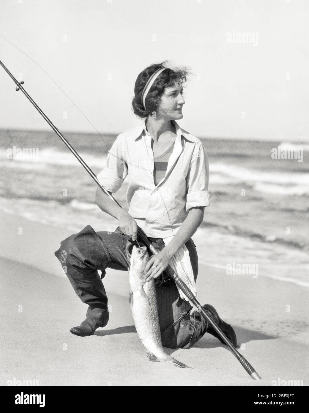 1920s WOMAN KNEELING ON OCEAN BEACH SHORE WITH ROD AND A WEAKFISH CAUGHT SURF  FISHING - a2545 HAR001 HARS CATCH LADIES PERSONS UNITED STATES OF AMERICA  ATHLETIC HOOK ROD SURF B&W NORTH