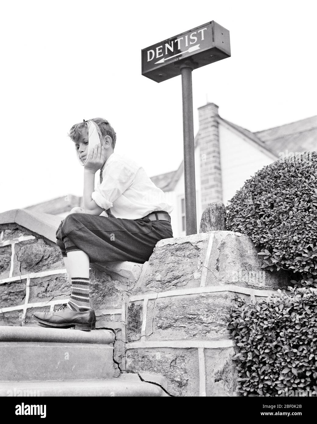 1930s 1940s BOY SITTING ON STONE WALL UNDER A SIGN POINTING TO DENTIST OFFICE HOLDING HIS FACE  SUFFERING TOOTHACHE  - b12235 HAR001 HARS B&W SADNESS DENTAL SUFFERING HIS LOW ANGLE AGONY DIRECTION TOOTHACHE IRRITATION CONCEPTUAL TENDERNESS DISCOMFORT GROWTH JUVENILES PAINFUL SOLUTIONS ACHE BLACK AND WHITE CAUCASIAN ETHNICITY HAR001 OLD FASHIONED Stock Photo