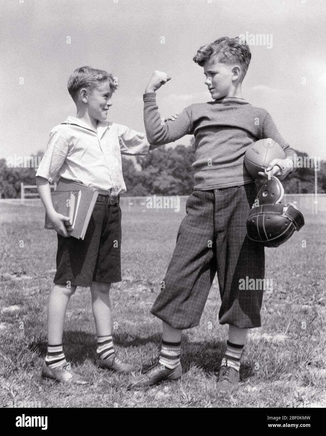 1930s  SCHOOLBOY FEELING ARM MUSCLE BICEP OF HIS OLDER STRONGER  FOOTBALL PLAYER BROTHER - b4463 HAR001 HARS HAPPINESS HIS STRENGTH RECREATION PRIDE BICEP FEELING SIBLING CONNECTION CONCEPTUAL FRIENDLY MUSCLES STYLISH COOPERATION GROWTH JUVENILES PRE-TEEN PRE-TEEN BOY TOGETHERNESS BLACK AND WHITE CAUCASIAN ETHNICITY HAR001 OLD FASHIONED Stock Photo