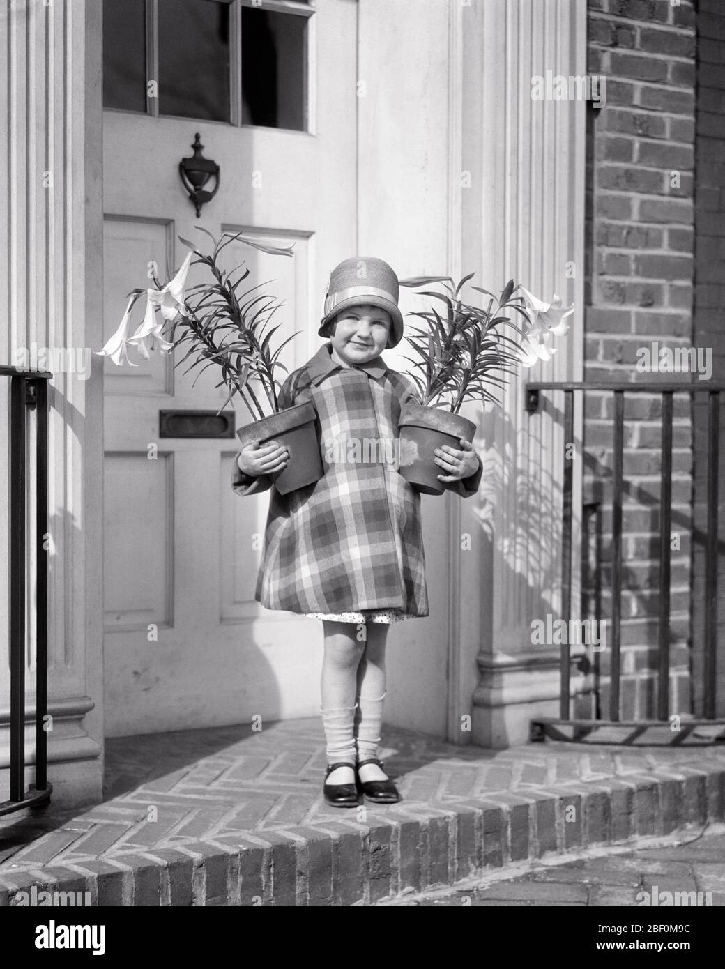 1920s GIRL WEARING CLOCHE HAT PLAID COAT MARY JANE SHOES SOCKS FALLING DOWN HOLDING TWO POTS OF EASTER LILIES AT FRONT DOOR - e89 HAR001 HARS JOY LIFESTYLE SATISFACTION RELIGION FEMALES EASTER HOME LIFE COPY SPACE FRIENDSHIP FULL-LENGTH INSPIRATION FESTIVAL CHRISTIAN PLAID CONFIDENCE MARY B&W EYE CONTACT CHEERFUL CHRISTIANITY EXTERIOR CLOCHE SMILES CONNECTION JOYFUL STYLISH LILIES JANE JUVENILES BLACK AND WHITE HAR001 OLD FASHIONED Stock Photo