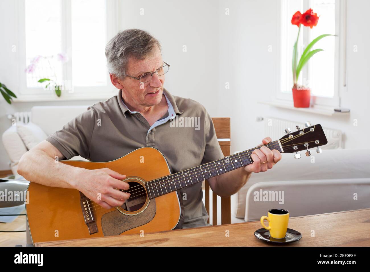 Senior man playing guitar at home in a bright living room with a cup of espresso on the table Stock Photo