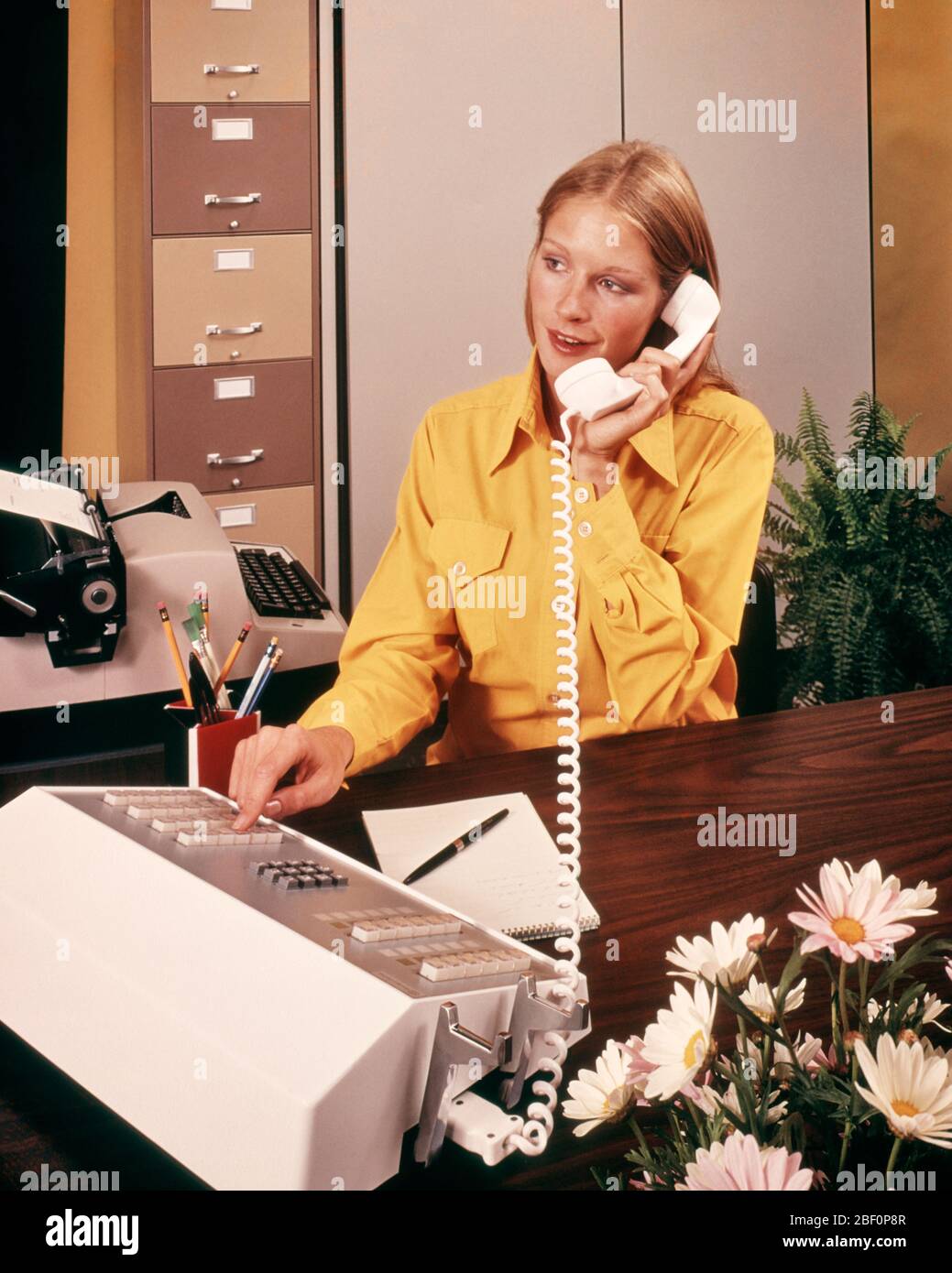 1970s BLONDE RECEPTIONIST YELLOW BLOUSE ANSWERING PHONE PRESSING BUTTON ON TELEPHONE SWITCHBOARD CONSOLE DAISY ARRANGEMENT  - ko900 PHT001 HARS LADIES PERSONS GROWN-UP CONFIDENCE ANSWERING DAISY WORK PLACE SWITCHBOARD CONSOLE HIGH ANGLE CUSTOMER SERVICE OFFICE WORKER BLOUSE OCCUPATIONS CONNECTION ELECTRIC APPLIANCE GAL FRIDAY ADMINISTRATOR SECRETARIES STYLISH PRESSING AMANUENSIS COMMUNICATE YOUNG ADULT WOMAN CAUCASIAN ETHNICITY CLERICAL OLD FASHIONED Stock Photo