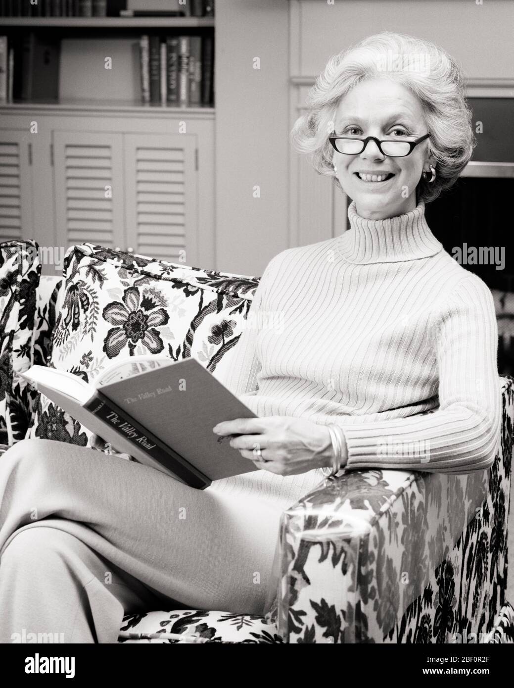 1970s MATURE RELAXED SLENDER HEALTHY WOMAN HOLDING A BOOK WEARING READER EYEGLASSES TURTLENECK SWEATER LOOKING AT CAMERA SMILING - r24219 HAR001 HARS PHYSICAL FITNESS PERSONS CONFIDENCE EYEGLASSES EXPRESSIONS MIDDLE-AGED B&W EYE CONTACT HAPPINESS MIDDLE-AGED WOMAN WELLNESS CHEERFUL LEISURE KNOWLEDGE RELAXED SLACKS SMILES WHITE HAIR CONTENT JOYFUL READER STYLISH RELAXATION SLENDER AT HOME BLACK AND WHITE CAUCASIAN ETHNICITY HAR001 OLD FASHIONED TURTLENECK Stock Photo