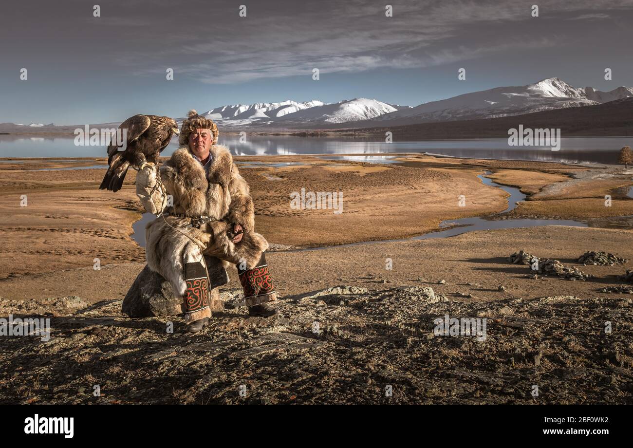 Mongolian eagle hunter posted with lake in front of landscape with lake and snow-covered mountains, Bajan-Oelgii province, Mongolia Stock Photo