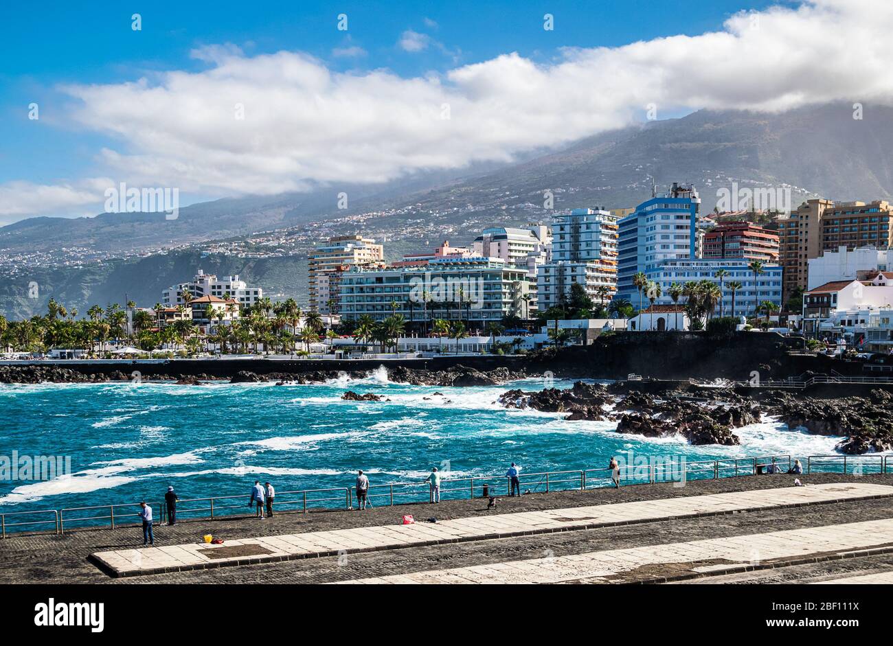 Locals fishing and watching giant waves crashing the shore of the port area on Puerto de la Cruz as the calima mist descends from mount Teide. Stock Photo
