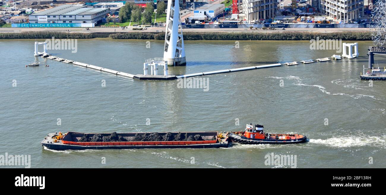 Aerial view River Thames tugboat pushing barge loaded heaps of excavated earth passing cable car pylon column & floating boom at North Greenwich UK Stock Photo