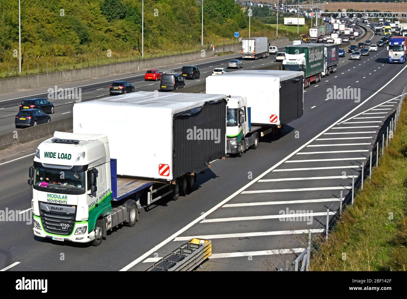 Looking down on slow moving traffic backing up on busy M25 motorway behind two lorry trucks loaded with wide load oversize modular building England UK Stock Photo