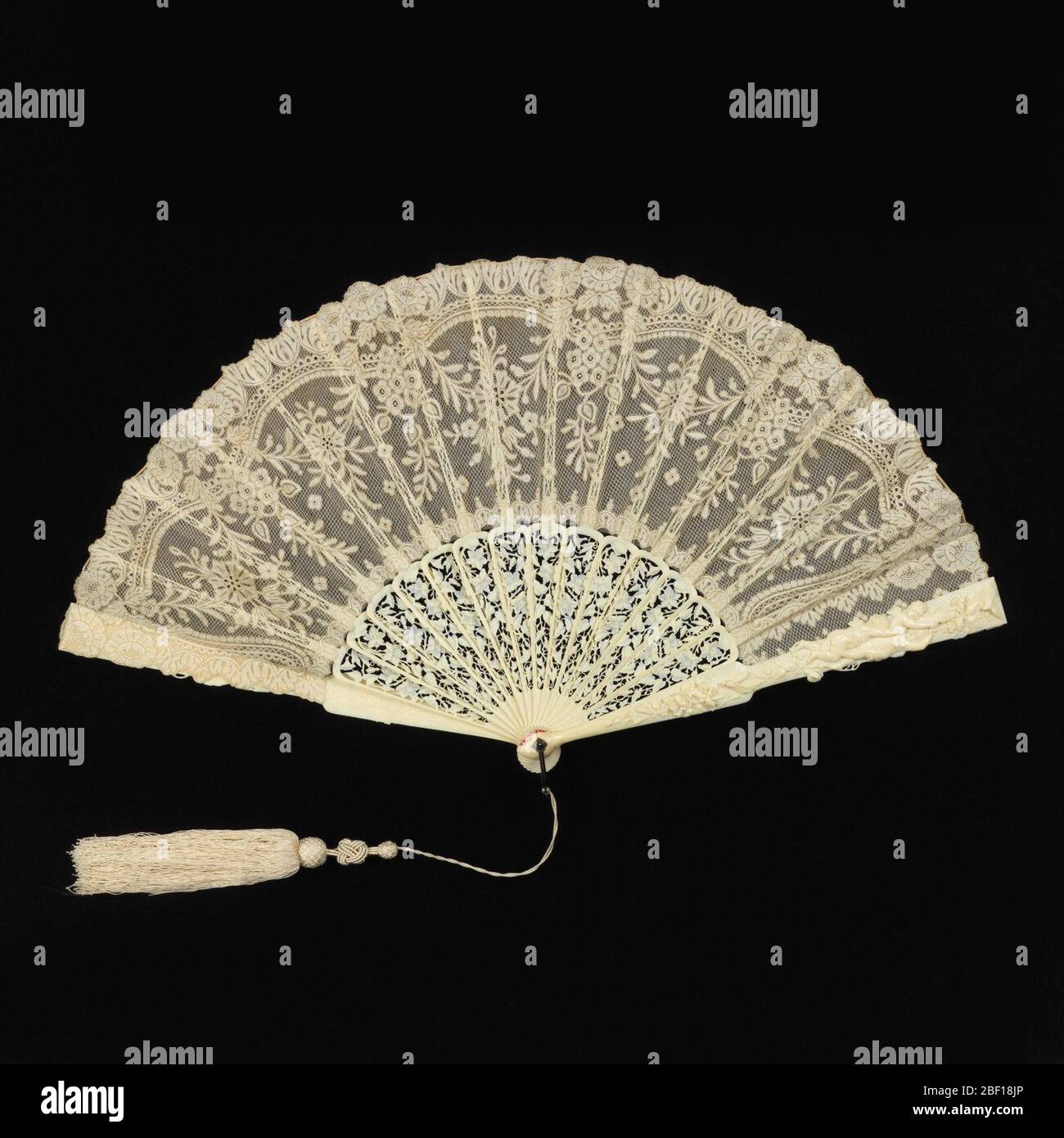 Pleated fan. Pleated fan. Leaf ofwhite cotton bobbin-made Valenciennes lace in floral design, backed with silk net. Sticks of pierced and carved ivory in floral design. Guards with figures of birds, cupids and insects. Metal loop at the rivet. Attached tassel. Stock Photo