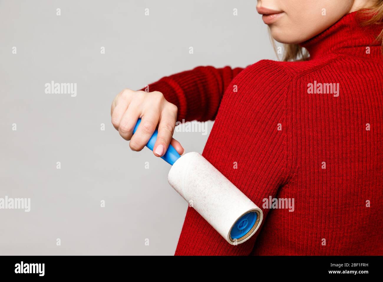 Woman hand using a sticky roller to clean fabrics - red woolen turtleneck from dust, hair, lint and fluff, front view, close up. Grey background. Stock Photo