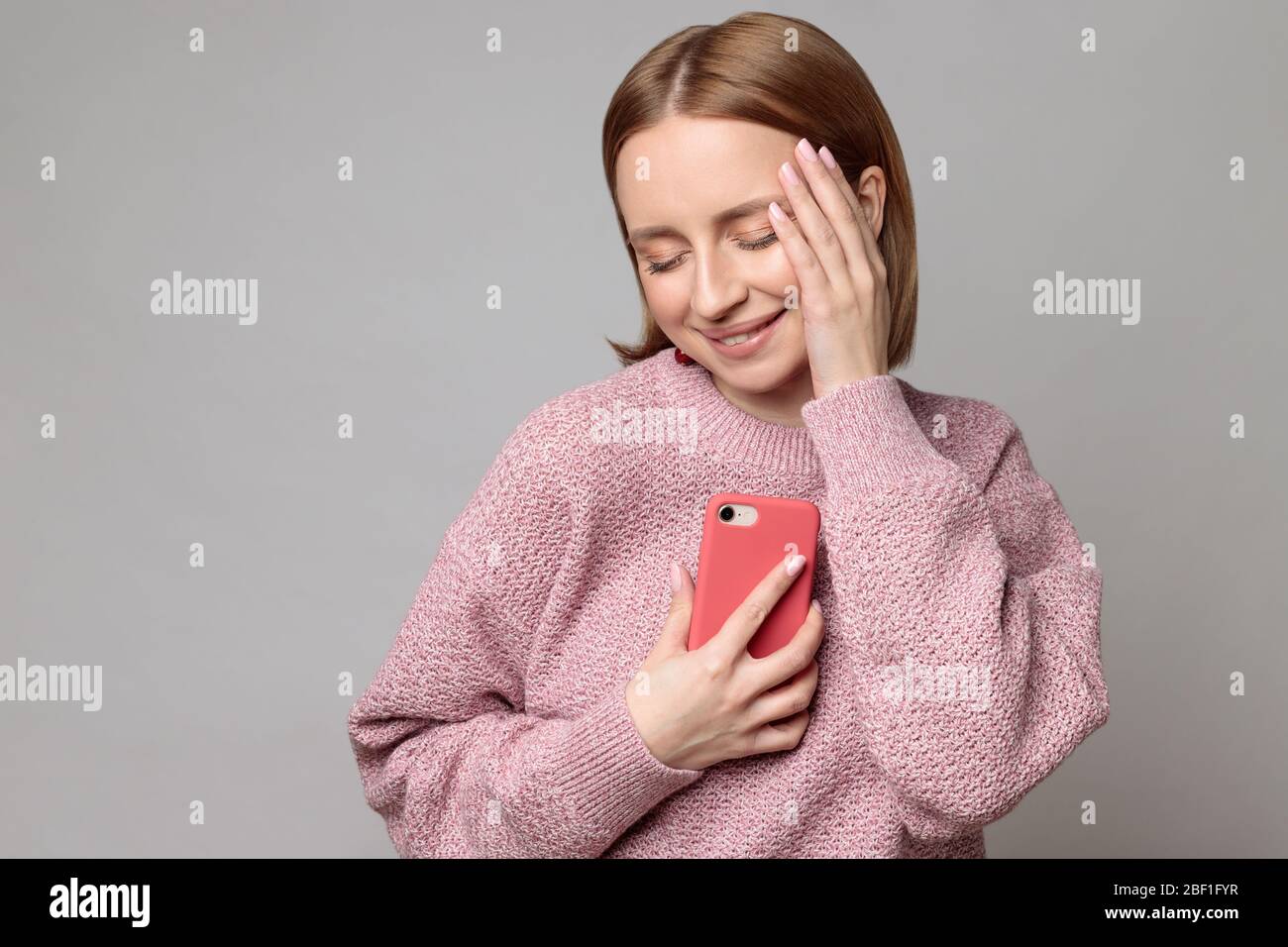 Oh so cute. Pleasant cheerful lovely european woman holding smartphone clasped to her chest and heart, closed eyes, isolated on grey background. Posit Stock Photo