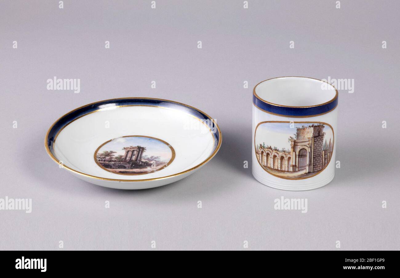 Cup and saucer. Short cylindrical cup (a) with loop handle; gray-white ground decorated with polychrome-painted oval scene showing classical ruin of building wall with arches; thin gilt border around oval; rim with underglaze blue band between two thin gilt lines; three incis Stock Photo
