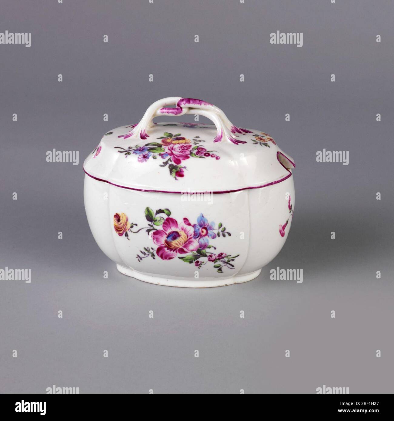 Sugar bowl lid and tray. Round bowl with large scalloped edges; lid has plant form loop handle. Lid rim is painted in deep purple. Decorated with deep pink-purple, yellow and blue blossoms and green leaves and stems on white background. Bowl's interior has painted green leaf. Stock Photo