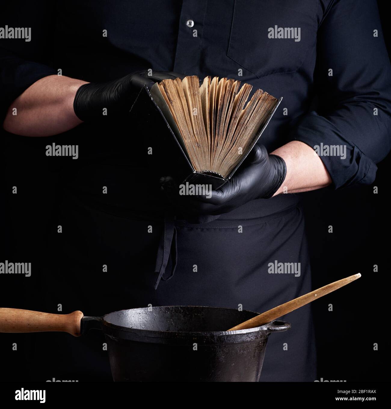 male cook in a black uniform holds an old cookbook with recipes, on a table is a cast-iron frying pan with a wooden handle, low key Stock Photo