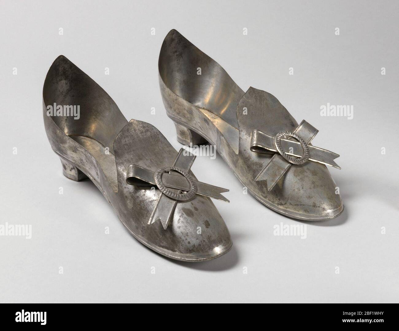 Pair of slippers. Pair of tin slippers on short heels; looped tin ribbons with nothced ends and stamped oval tin buckles mounted at tongues. Each heel, instep, back and sole made separately, soldered together. Stock Photo