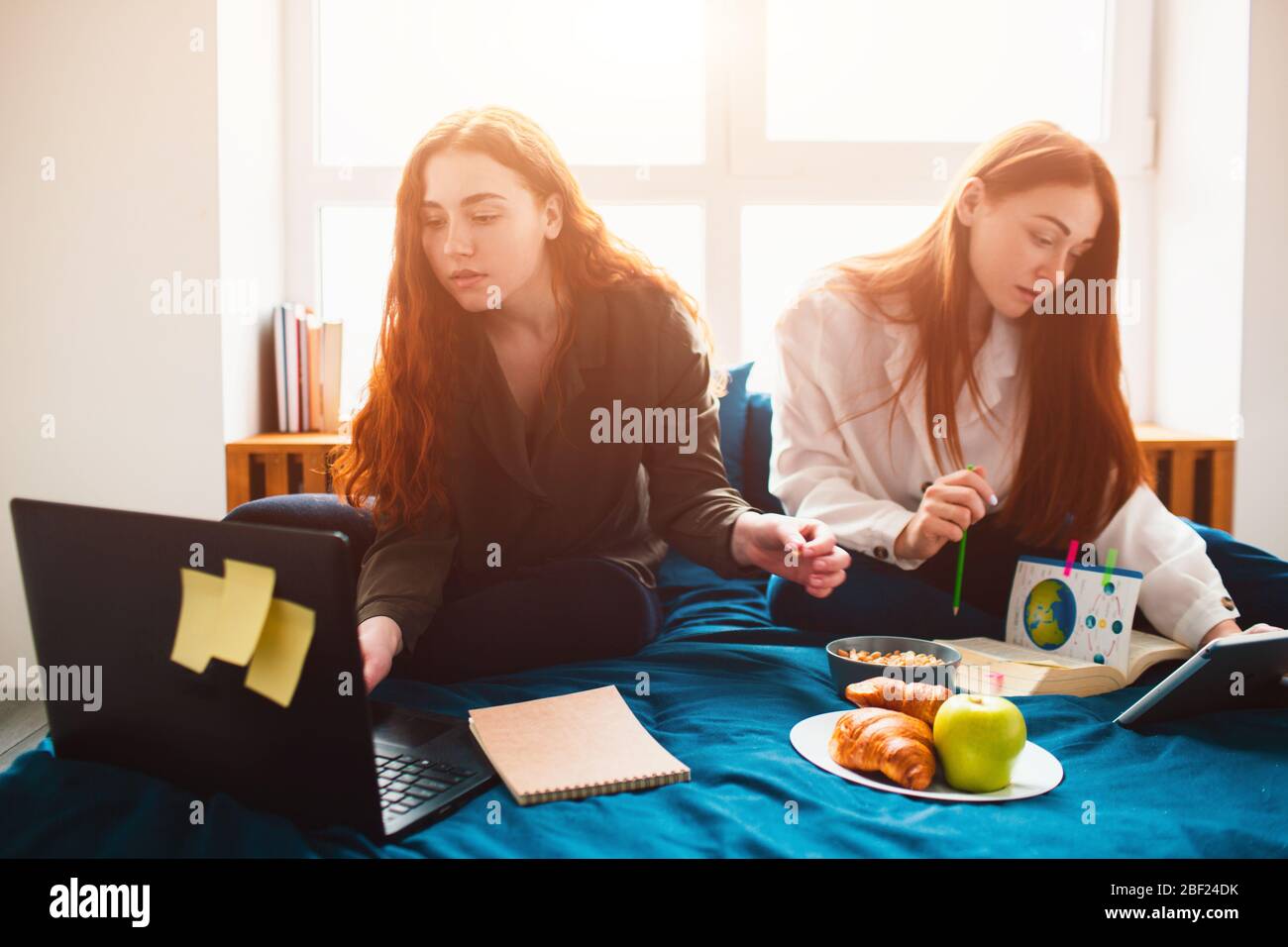 Two red-haired students study at home or prepare for exams. Young women doing homework in a dormitory bed near the window. There are notebooks, food Stock Photo