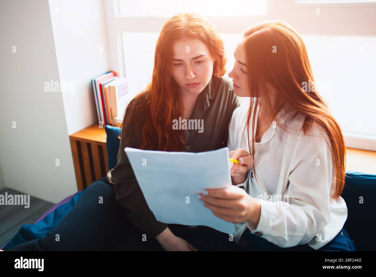 One student is holding a form or document or tests in her hands and the second is looking attentively. Two red-haired students study at home or in a Stock Photo